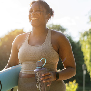 Woman with yoga mat and water bottle smiling outside
